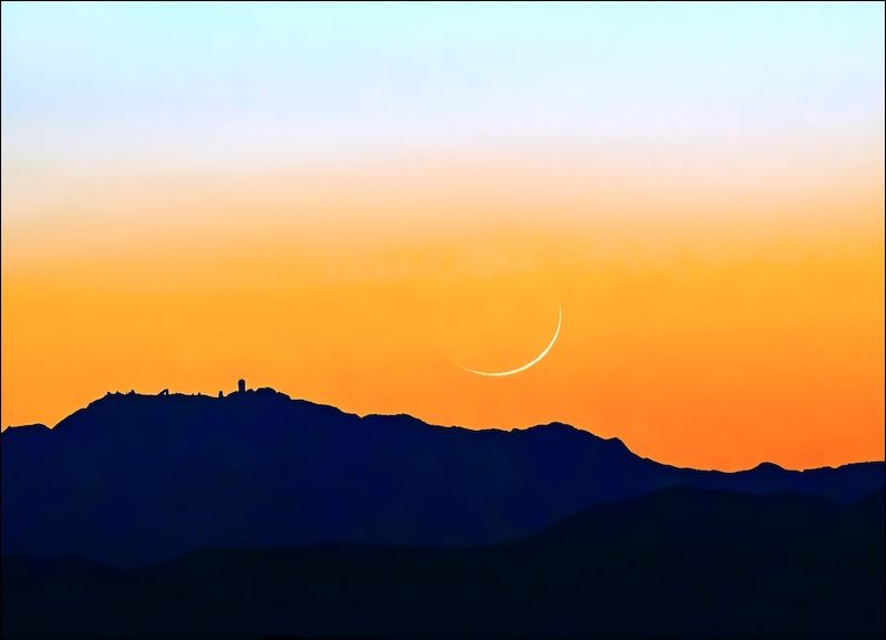 Dark foreground hills, orange sky fading to yellow and blue above, with a sliver of a moon.