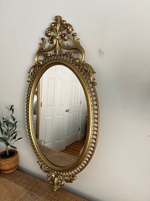 May include: A gold oval mirror with an ornate, scrolled frame. The frame has a beaded design around the perimeter and a large, intricate scroll design at the top and bottom.