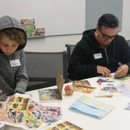 A father and son cut out papers and work on a craft project together while sitting at a table.