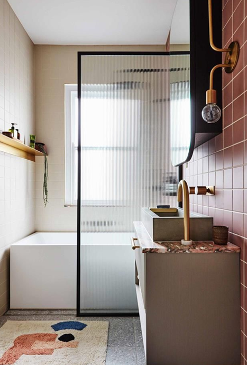 Modern bathroom with a glass shower partition, a rectangular tub, beige and pink tiled walls, a floating shelf with plants, a patterned rug, a vessel sink, and a gold faucet with an exposed bulb light fixture.