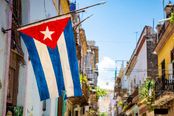 The image presents a vibrant street scene in Cuba, prominently featuring the Cuban flag hanging from a pole on the left, which immediately establishes the location. The narrow street, flanked by weathered colonial buildings with balconies and shutters, is a distinctive and recognizable element of Cuban urban life. The bright blue sky and strong sunlight contribute to a warm, inviting atmosphere, enhancing the image's appeal. The composition is well-framed, with the flag drawing the viewer's eye into the depth of the street, creating a sense of exploration. The horizon is level, and the framing is balanced, avoiding awkward cropping or excessive sky. There are minimal distractions; a few utility poles and wires are present but do not detract significantly. The lighting is bright and natural, with good color saturation that captures the vivid blues and whites of the flag and the warm tones of the buildings. The image is sharp and clear, with no noticeable motion blur, noise, or focus issues. There are no people in the foreground to dominate the scene, and no text overlays or borders. The overall beauty is high, with a strong sense of place and cultural identity. This image effectively conveys the charm and character of Cuba, making it highly suitable for a travel guide cover.