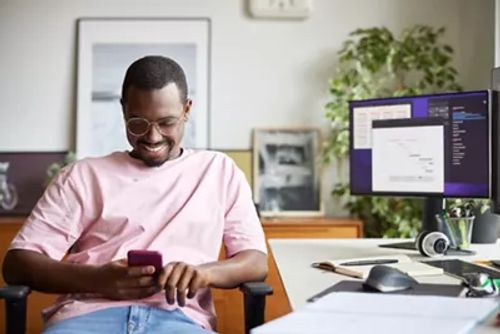 微笑的商人坐在电脑桌旁使用智能手机。