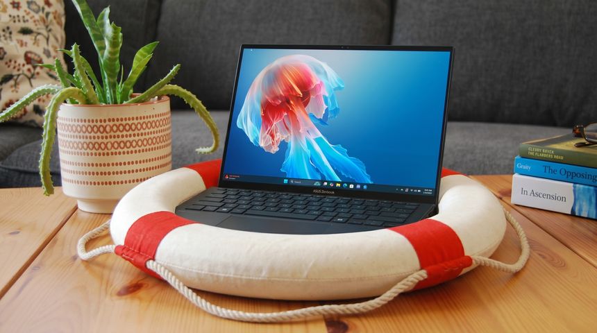 A laptop displaying a colorful jellyfish screensaver rests on a life preserver. A potted plant and stacked books add a cozy feel on the wooden table.
