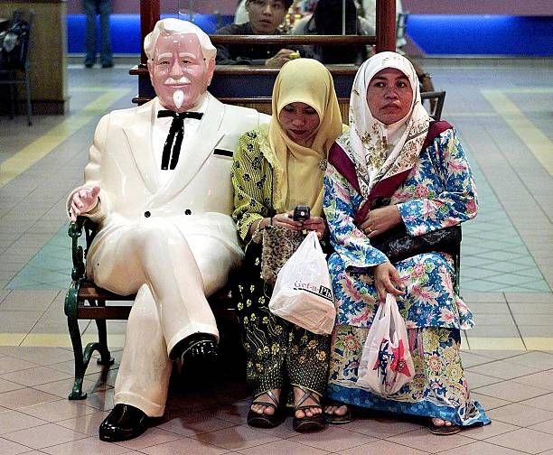 Two Malaysian women sit next to Colonel Sanders, the famous icon of Kentucky Fried Chicken , on a bench at a KFC outlet in Kuala Lumpur 18 July 2001....