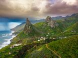 This image captures a dramatic and picturesque landscape of the Canary Islands, featuring iconic volcanic peaks, lush green slopes, a winding coastal road, and a rugged shoreline under a moody, cloud-filled sky. The scene is highly recognizable as the Canary Islands, particularly resembling locations like La Gomera or Tenerife, with strong match confidence. The composition is well-framed, with the prominent rock formations serving as natural focal points, and the horizon is level, contributing to a balanced and visually appealing layout. The lighting is dynamic, with strong contrast between the dark clouds and the illuminated green hills, enhancing the scene's drama and beauty. There are no visible distractions such as poles, vehicles, or people, and the image is free from unwanted elements like text overlays or borders. The technical quality is high, with sharp detail in the landscape and no noticeable noise or motion blur. The overall beauty of the scene is exceptional, making it highly suitable for a travel guide cover. The image effectively conveys the natural splendor and adventurous appeal of the Canary Islands.