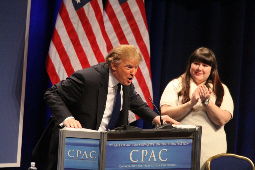 Trump, leaning heavily onto a lectern, with his mouth open mid-speech and a woman clapping politely next to him