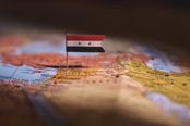 The image features a miniature Syrian flag placed on a map, with Syria's location highlighted. The composition uses a shallow depth of field, focusing sharply on the flag and the immediate area of the map, while the background and foreground are blurred. This creates a strong focal point and a sense of place. The color palette is warm and earthy, with the red, white, and black of the flag providing a striking contrast against the map's colors. The lighting is soft and even, enhancing the textures of the map and the flag. There are no distracting elements such as poles, vehicles, or hands; the frame is well-balanced, with the subject centrally positioned. The image is technically sound, with sharp focus on the intended subject and no visible noise or artifacts. The overall aesthetic is clean and evocative, suitable for a travel guide cover. However, the image is a stock photo with a watermark, which is a significant issue for commercial use. The watermark is prominent and would need to be removed. Despite this, the visual elements are strong and the image effectively conveys the theme of travel to Syria. The match confidence to 'syria' is high due to the clear presence of the Syrian flag and the map's geographical context. The distinctive features are the flag and the map, which are easily recognizable. The composition is well-executed, with no awkward cropping or tilted horizon. The lighting is appropriate, and the technical quality is good. There are no unwanted elements like borders or text overlays, and no people are present. The overall beauty of the image is high, making it suitable for a travel guide cover, but the watermark detracts from its final suitability.