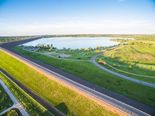 This aerial image of Cherry Creek presents a scenic view of a large reservoir bordered by green parkland and a prominent dam structure. The composition is well-framed, with the curved dam and pathways leading the eye toward the water and the distant shoreline. The horizon is level, and the framing avoids awkward cropping, with the expansive sky contributing to a sense of openness. The lighting is bright and even, with warm golden tones suggesting late afternoon, enhancing the natural beauty of the landscape. There are no visible distractions such as vehicles, poles, or people, and the image is free of text overlays or unwanted elements. The technical quality is high, with sharp focus, minimal noise, and vibrant colors. The scene is highly recognizable as a recreational area, with distinctive features like the dam and winding paths contributing to its identity. The overall aesthetic is tranquil and inviting, making it an excellent candidate for a travel guide cover. The match confidence to 'Cherry Creek' is high due to the recognizable landscape features and the name's association with the area.
