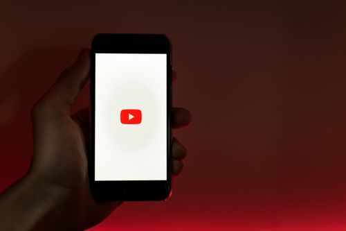 Free A hand holding a smartphone displaying the YouTube app against a red background. Stock Photo