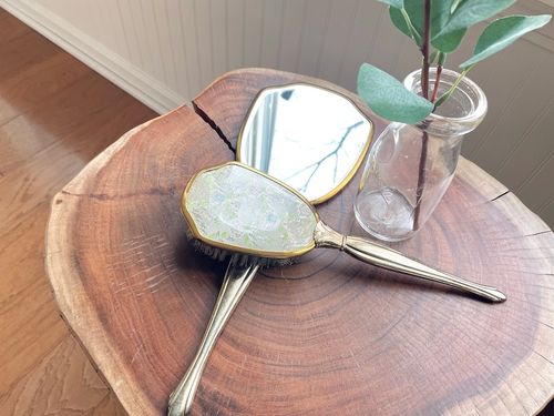 May include: A vintage hand mirror and hairbrush set with gold trim. The mirror is oval shaped and the brush has a clear handle with a floral design. The set is on a wooden surface.