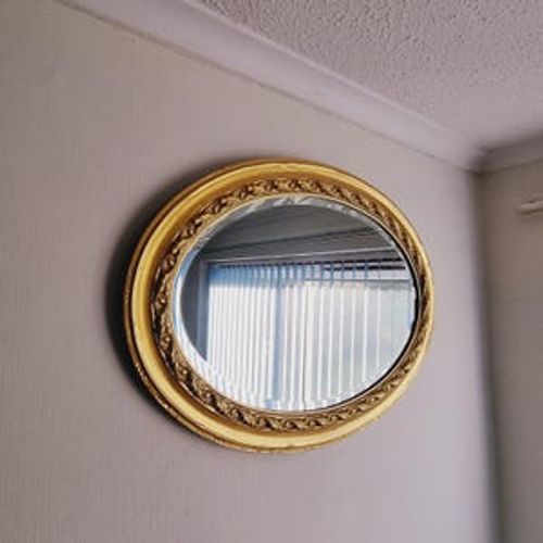 May include: A gold oval mirror with a decorative ornate frame. The mirror reflects a window with white blinds.