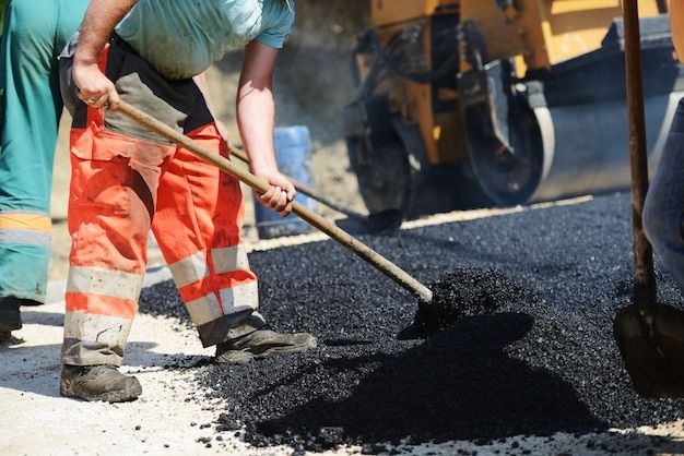 Two construction workers in high-visibility vests and hard hats are operating a large road paver machine. They are laying fresh asphalt on a road, with one worker standing on the machine and the other standing on the ground beside it. The scene is set outdoors with some trees and a wooden fence in the background.