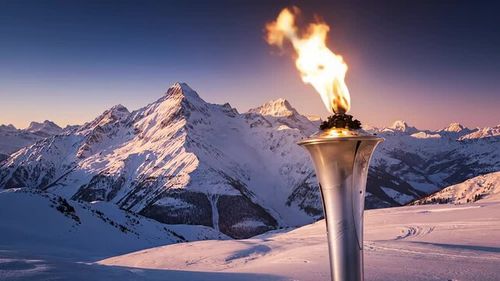 The fiery Olympic torch with a radiant flame representing the spirit of global athletic competition. Majestic snowy alpine mountains in the distance creating a stunning backdrop