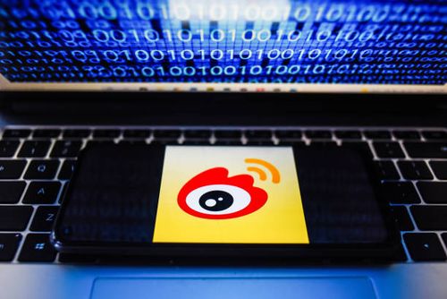 In this photo illustration a Weibo logo seen displayed on a smartphone with binary code on a laptop screen.