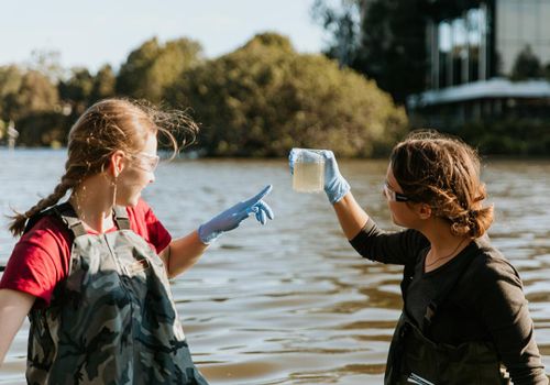 Students stand in a lake looking at a water sample in a beaker