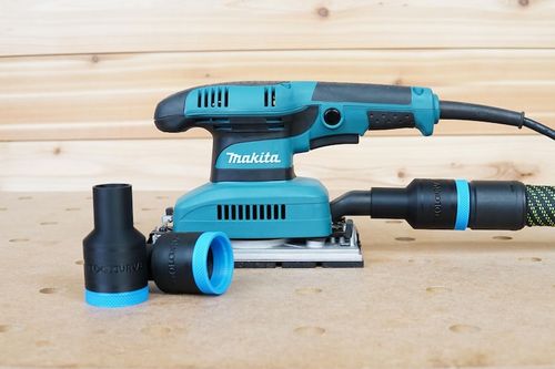 May include: A teal Makita random orbital sander with a dust collection system. The sander has a comfortable grip and a large pad for efficient sanding.  Two black dust collection adapters with blue accents are also shown. The brand name Makita is visible on the sander. The tool is displayed on a light brown wooden surface.