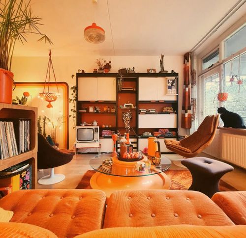 Bright retro 70s living room featuring an orange sofa, low coffee table, wall unit with TV and playful accessories.