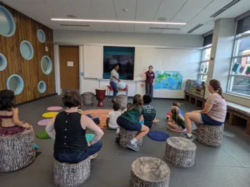 Kids and families sit on imitation wood poofs as someone leads an international storytime at Pinney Library