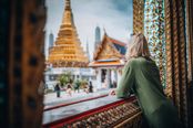 This image captures a scenic view of a golden temple in Thailand, likely the Grand Palace in Bangkok, seen through an ornate window frame. The composition is strong, with the window acting as a natural frame that draws the eye to the iconic architecture. The subject is well-balanced, with the woman in the foreground providing a sense of scale and perspective, while the temple remains the clear focal point. The lighting is soft and even, enhancing the warm golden tones of the temple and the vibrant colors of the window. The image is sharp and free of motion blur or noise, with good focus on the temple in the background. There are no distracting elements such as poles, vehicles, or clutter; the people in the distance are small and do not dominate. The horizon is level, and the framing is appropriate, with no awkward cropping or cut-off subjects. The overall beauty of the scene is high, with a harmonious blend of cultural richness and aesthetic appeal. The image effectively conveys the essence of Thailand, making it highly suitable for a travel guide cover. The match confidence to 'Thailand' is high due to the distinctive and recognizable architecture of the temple and the traditional window design. The presence of people is minimal and does not detract from the scene, adding a human element without overwhelming it. Overall, the image is technically sound and visually compelling.