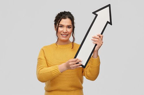 人们的概念 - 一位快乐微笑的年轻女性，鼻子上有穿孔，手持一支大白色粗箭头，指向上方，背景为灰色。图片下载