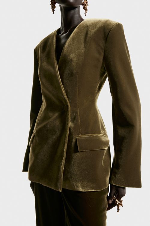 View larger image: An olive green velvet blazer is being worn by the model, featuring a wrapover front with a covered button, structured shoulder pads, and a nipped-in silhouette from waist darts. A jetted flap pocket is visible on the front, and the blazer is being styled with matching velvet pants.