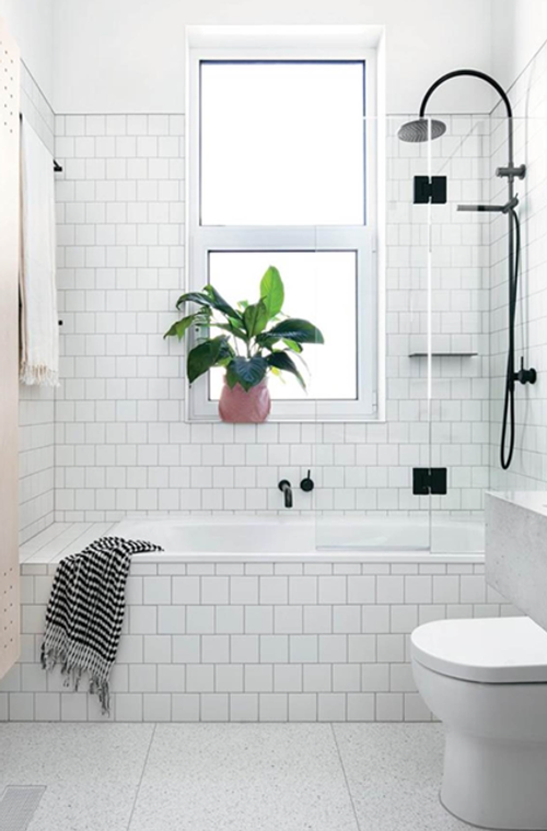 Modern bathroom with white subway tiles, a bathtub, black fixtures, a glass shower screen, a toilet, a plant hanging in front of the window, and a black-and-white striped towel draped over the tub.