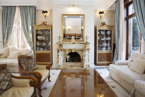 Sophisticated living room features a classic fireplace designed with Greek sculptures and gold mirror situated between display cabinets lighted by wall sconces.