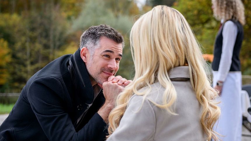 Yannik und Larissa sitzen im Freien. Er schaut sie verliebt an und hält dabei die Hände verschränkt an sein Kinn.