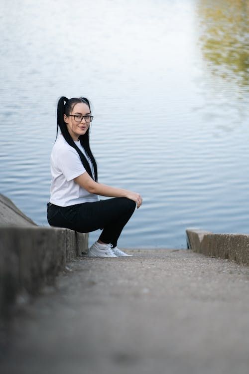 Free A young woman with long hair and glasses poses by a calm riverside, exuding a relaxed vibe. Stock Photo