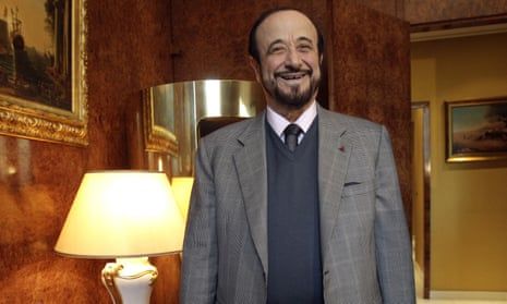 Rifaat al-Assad wears a grey suit and stands in an opulent Paris apartment with paintings on the wall