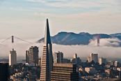 This image of San Francisco presents a classic and recognizable cityscape, featuring the Transamerica Pyramid prominently in the foreground, with the Golden Gate Bridge partially obscured by fog and distant hills in the background. The match confidence to San Francisco is excellent due to the unmistakable skyline and iconic landmarks. The composition is well-framed, with the pyramid centered and the bridge and mountains providing depth and context; the horizon is level, and the framing includes the essential elements without awkward cropping. The fog adds a dramatic, atmospheric quality, enhancing the city's mystique. The lighting is soft and diffused, typical of a foggy morning, creating a moody and appealing ambiance with balanced colors. The technical quality is high, with sharp focus on the central structures and no visible motion blur or noise. There are no distracting elements such as poles, vehicles, or people; the image is clean and uncluttered. The overall beauty is strong, capturing the essence of San Francisco's unique character. The image is highly suitable for a travel guide cover, effectively conveying the city's iconic appeal and scenic beauty.