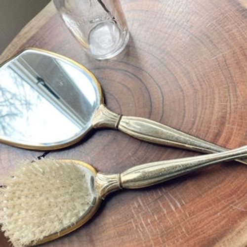 May include: A vintage silver-toned hand mirror and hairbrush set with gold accents. The mirror has a rounded oval shape and the brush has a rectangular shape. Both have ornate handles.