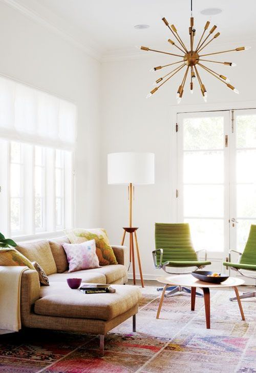 A mid-century modern living room with a bold printed rug, a sectional sofa with bright pillows, green chairs and a coffee table, a burst chandelier.