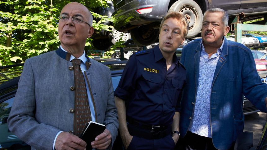 "Die Rosenheim-Cops - Der Schrottkönig": Polizeidirektor Achtziger (Alexander Duda), Polizeihauptmeister Mohr (Max Müller ) und Kommissar Hofer (Joseph Hannesschläger) stehen nebeneinander vor einer Reihe schrottreifer Autos und schauen in die gleiche Richtung. Achtziger hält sein Notizbuch in den Händen.