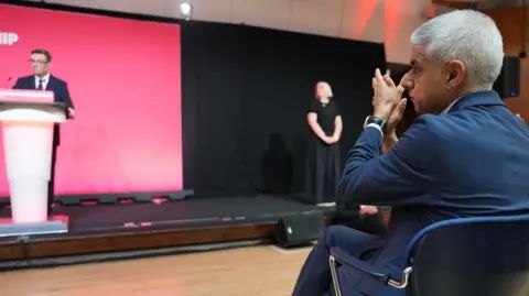 A seated Sir Sadiq Khan applauds Andy Burnham, who is standing at a podium in front of a red backdrop.
