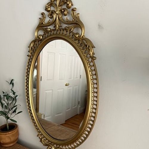May include: A gold oval mirror with an ornate, scrolled frame. The frame has a beaded design around the perimeter and a large, intricate scroll design at the top and bottom.
