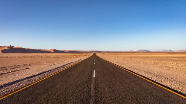 Open Desert Road through Sossusvlei Desert in Namibia Empty Open Desert Road in arid vast Desert Landscape of the Sossusvlei Namib Desert of Namibia. Giant Sand Dunes roadside the Highway in the background. Roadside panorama under blue summer skyscape. Namibian Desert, Sossusvlei, Namibia, Southern Africa. wide open highway stock pictures, royalty-free photos & images