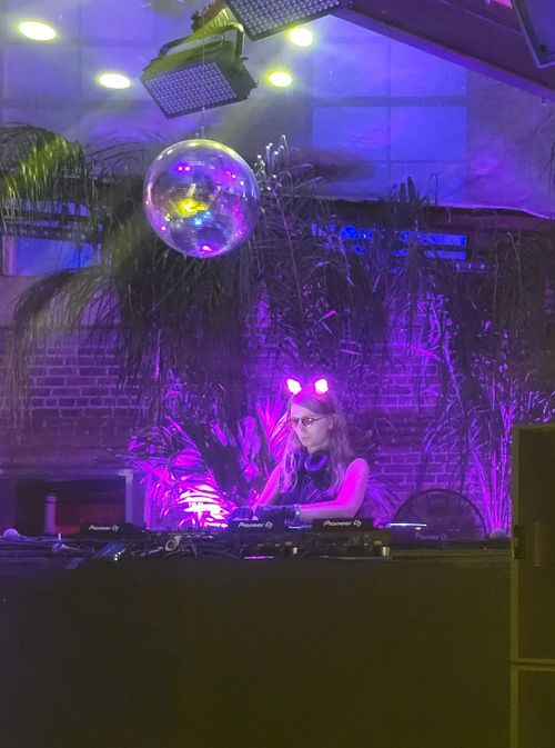 Chelsea Manning, wearing glowing cat ears, working behind a DJ booth with a discoball overhead