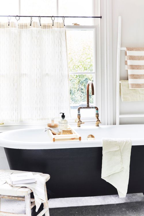 a small bathroom painted white with sheer white curtains and a black and white freestanding bath tub