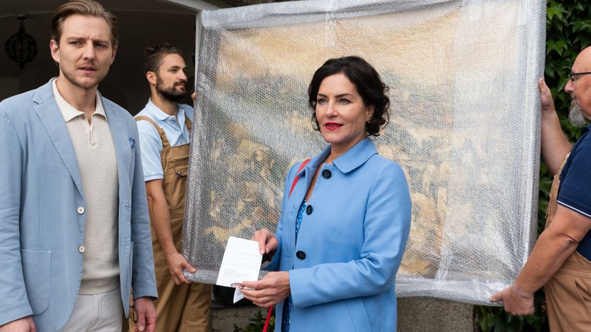 "Die Rosenheim-Cops - Stockls Verdacht": Thomas Lob (Matthias Zera) und Miriam Stockl (Marisa Burger) stehen nebeneinander und schauen in dieselbe Richtung. Hinter ihnen tragen zwei Mitarbeiter eines Transportunternehmens ein Gemälde aus dem Haus.