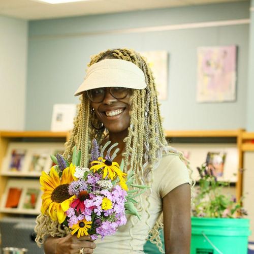 A black woman stands with an arrangement of flowers wearing a visor and patchwork jean shorts smiling in a library