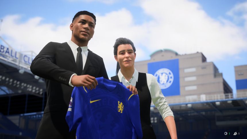 Young footballer in a black suit holds up a Chelsea home shirt beside a club representative during a signing presentation at Stamford Bridge, with Chelsea branding visible in the background.
