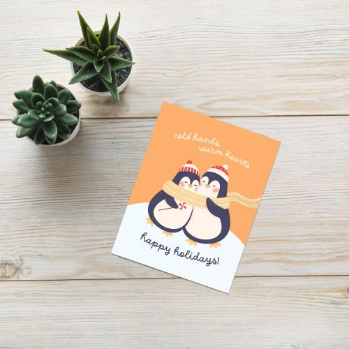 unique holiday card with two penguins hugging, wearing scarves and hats, on an orange background with text "Two penguins in knit hats sharing a scarf; text “cold hands, warm hearts — happy holidays!”"