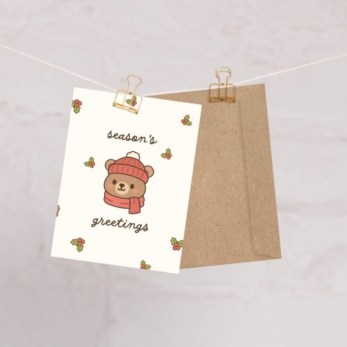 cute Christmas card with a bear wearing a pink hat and scarf, surrounded by holly leaves and berries with text: season's greetings