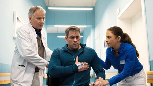 Astronaut Adam Birken (Marc Barthel, M.) sitzt in einem Rollstuhl und hält sich an die Brust. Dr. Roland Heilmann (Thomas Rühmann, li.) und Arzu Ritter (Arzu Bazman) kümmern sich um ihn.