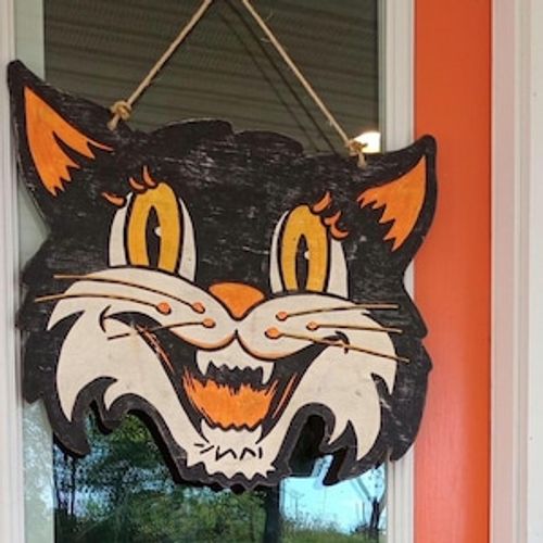 May include: A vintage-style Halloween decoration featuring a black cat face with orange ears, eyes, and mouth. The cat has a wide, toothy grin and is suspended by a natural twine rope. The wooden sign is distressed, giving it a rustic appearance.