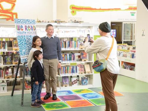 A family stands in front of a poster board and easel while another person snaps a picture of them inside a library