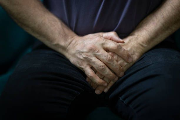 man sitting on sofa clutching his groin in pain - erectile dysfunction stock pictures, royalty-free photos & images