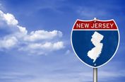 The image features a New Jersey state highway sign against a bright blue sky with scattered white clouds. The sign is clearly labeled 'NEW JERSEY' with the state's silhouette, making it highly recognizable and directly relevant to the theme. The composition is well-framed, with the sign positioned to the right, leaving ample negative space on the left, which creates a balanced and visually appealing layout. The horizon is level, and there is no awkward cropping or tilted perspective. The lighting is bright and natural, with good color balance, enhancing the vivid blue of the sky and the red and white of the sign. The image is sharp and free from motion blur or noise, with no distracting elements such as vehicles, people, or poles in the frame. There are no borders, text overlays, or unwanted artifacts. The overall aesthetic is clean, professional, and inviting, capturing a sense of travel and discovery. The image effectively conveys the identity of New Jersey and would make an excellent front cover for a travel guide.