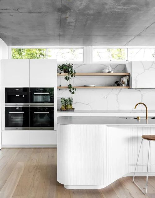 Modern kitchen with white cabinetry, marble backsplash, built-in ovens, gold faucet, floating shelves, and plants.